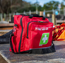 Load image into Gallery viewer, Red first aid kit with a green cross symbol on a wooden surface outdoors.