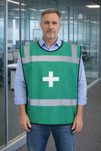 Load image into Gallery viewer, Man wearing a green safety tabbard style vest with a 'First Aid Cross' in an office setting